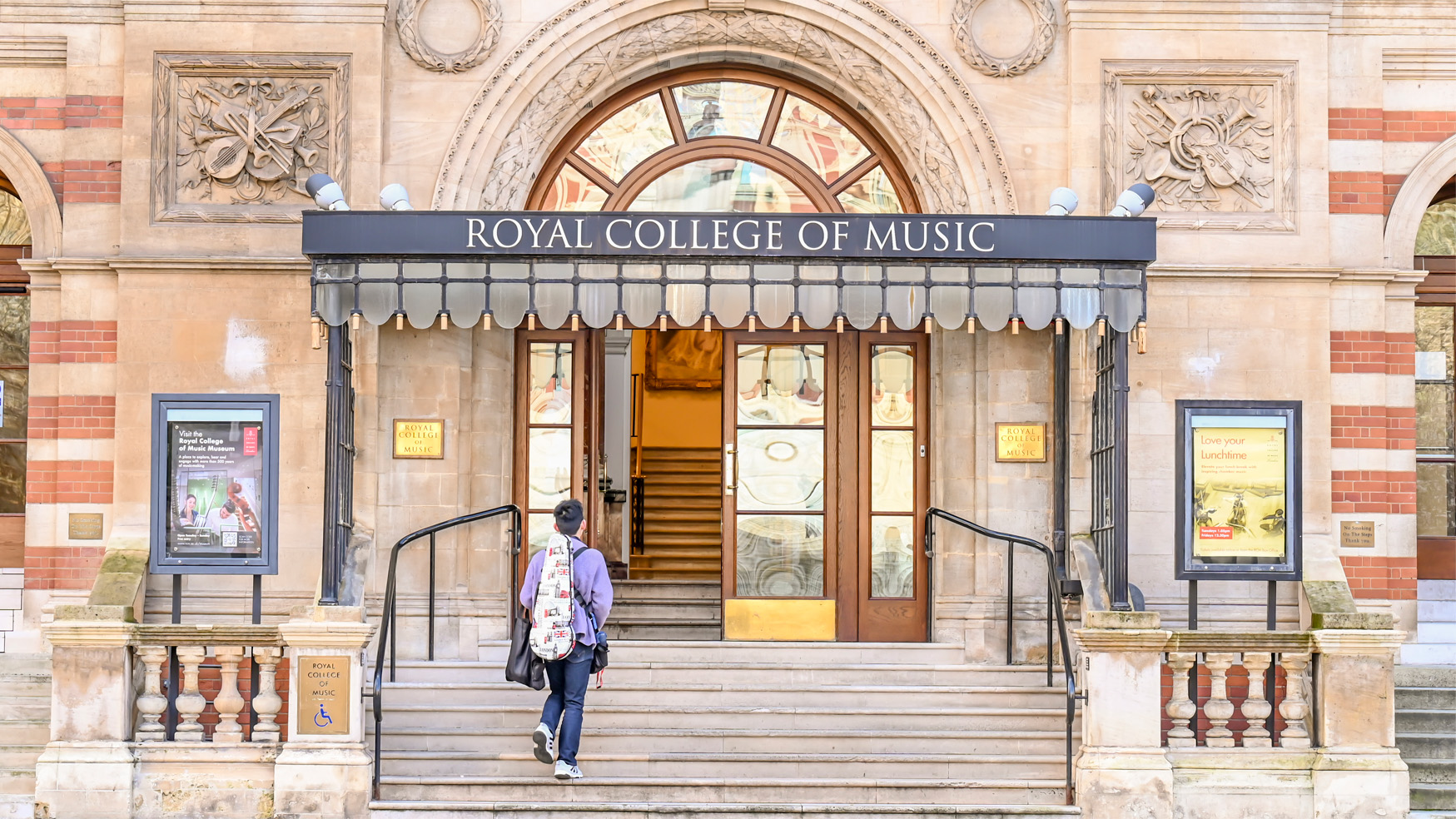 A student with a instrument case walking up the steps towards an open door, with the Royal College of Music sign above it